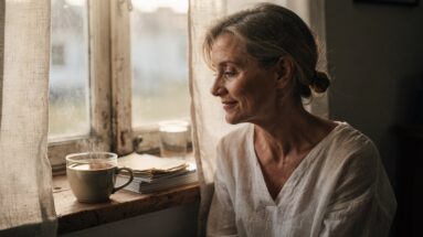 Mulher por volta dos quarenta e muitos anos, em perfil suave junto a uma janela com luz natural de manha, num momento tranquilo, com uma chavena de cha a fumegar num parapeito de madeira.